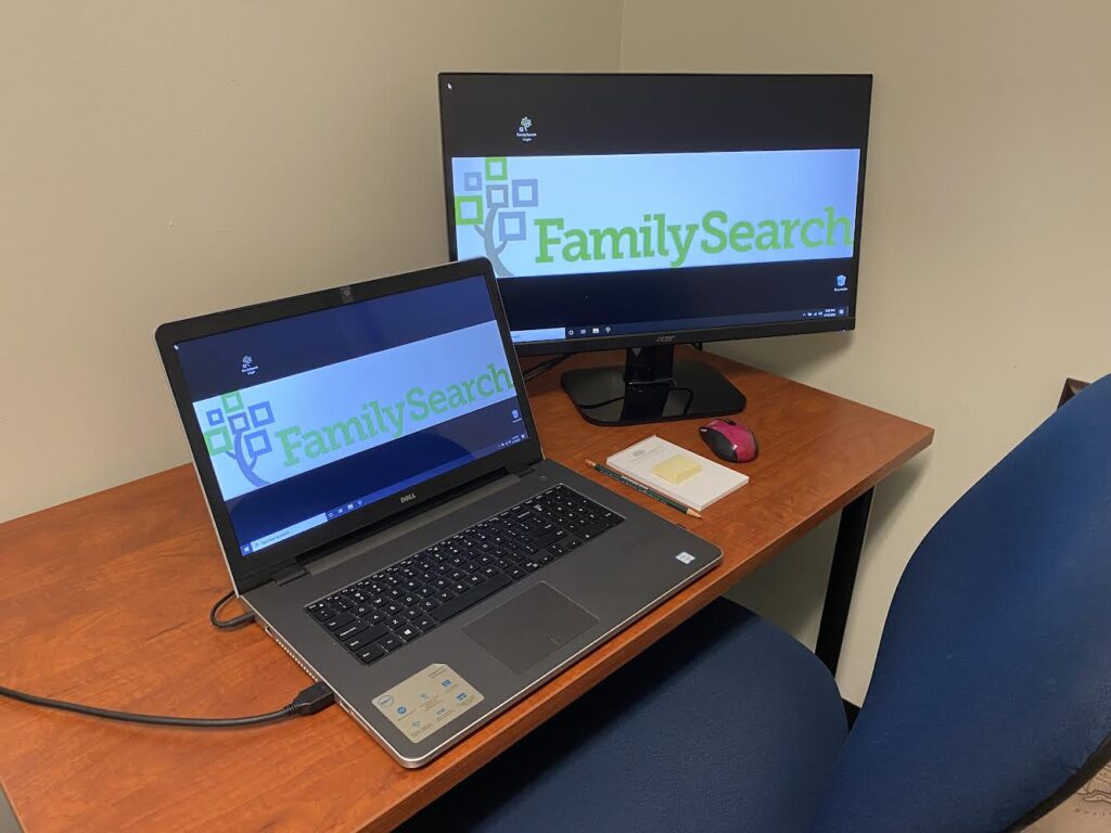 Laptop open with FamilySearch logo on screen and large monitor with FamilySearch logo on screen